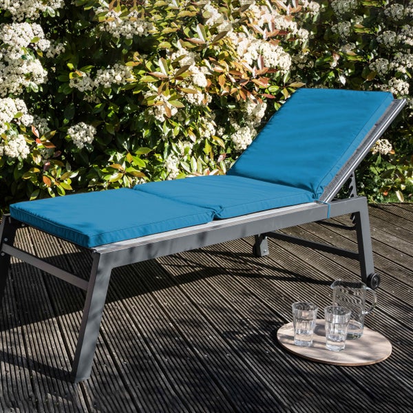 Sonnenliege mit grauem Metallgestell und petrolfarbener Polsterauflage auf einer Holzterrasse vor einer Hecke.