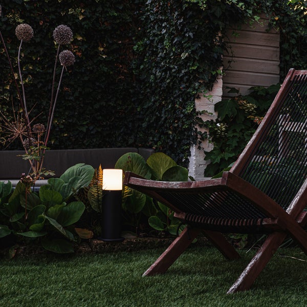 Schwarze Außenleuchte mit warmweißem Licht in einem Gartenbeet neben einem hölzernen Liegestuhl.