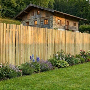 Hoher Sichtschutzzaun aus Holz im Garten mit Blumenbeet davor und rustikalem Haus im Hintergrund.