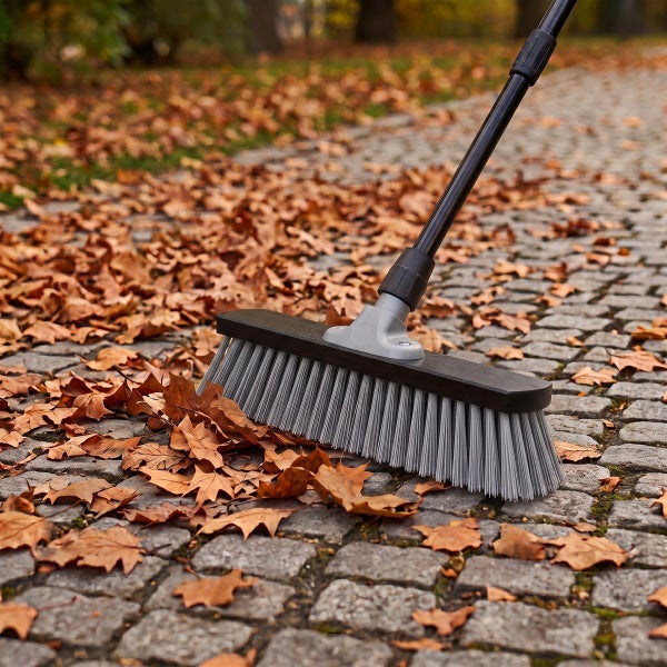 Ein Straßenbesen mit verstellbarem Teleskopstiel fegt trockenes Herbstlaub von einem gepflasterten Gartenweg.