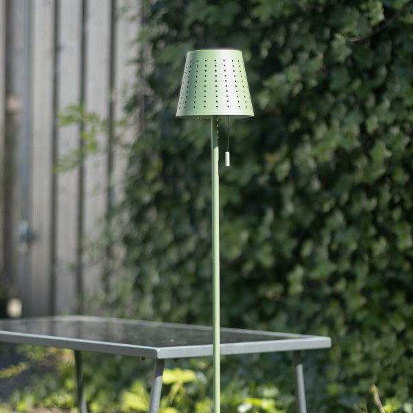 Grüne Stehlampe für den Außenbereich mit perforiertem Schirm und Zugschalter im Garten neben einem Tisch.