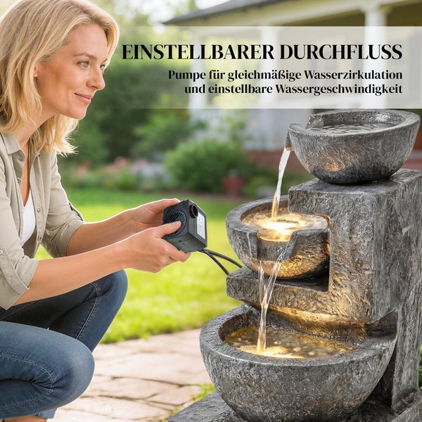Frau hält eine Pumpe neben einem dreistufigen Kaskadenbrunnen in Steinoptik mit Beleuchtung und fließendem Wasser im Garten.