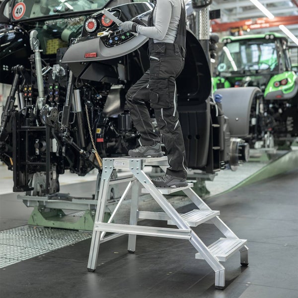 Eine Person nutzt eine Aluminium-Montagetreppe mit vier Stufen in einer Werkstatt, um Wartungsarbeiten an einer großen Maschine durchzuführen.