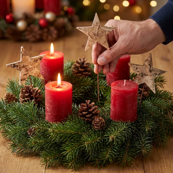 Hand platziert einen Stern aus Birkenrinde auf einem Adventskranz mit roten Stumpenkerzen und Tannenzapfen auf einem Holztisch.