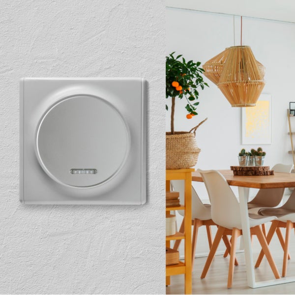 Ein moderner Wandschalter mit runder Wippe und Kontrollleuchte auf einer strukturierten Wand neben einer hellen Esszimmereinrichtung.