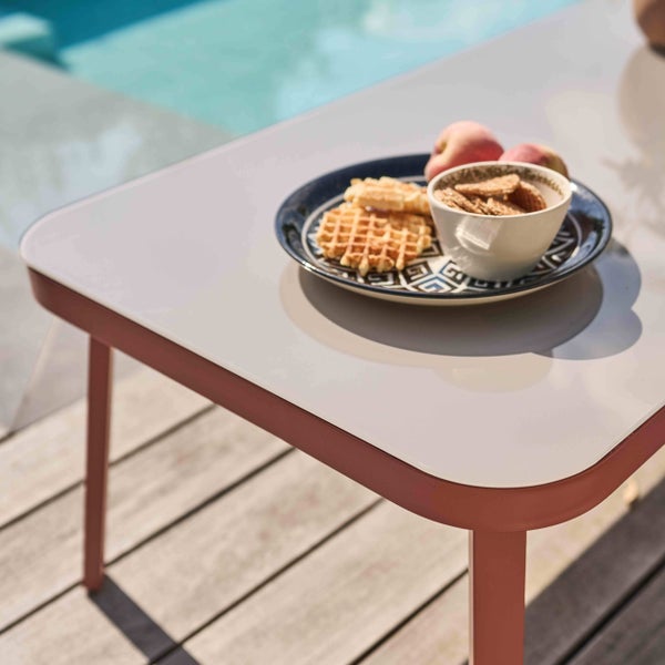Moderner Gartentisch mit hellen Tischplatte und terracottafarbenem Gestell auf einer Holzterrasse am Pool.