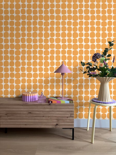 Modernes Zimmer mit orange gemusterter Tapete, einem Sideboard aus Holz, einer rosa Tischlampe und einer Vase mit Blumen.