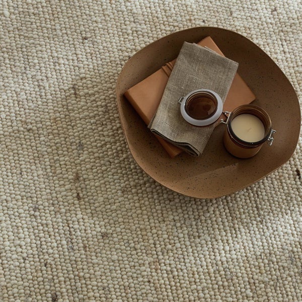 Detailansicht eines Teppichs mit Dekorationen, darunter ein Buch, eine Stoffserviette und eine Kerze in einem braunen Behälter.