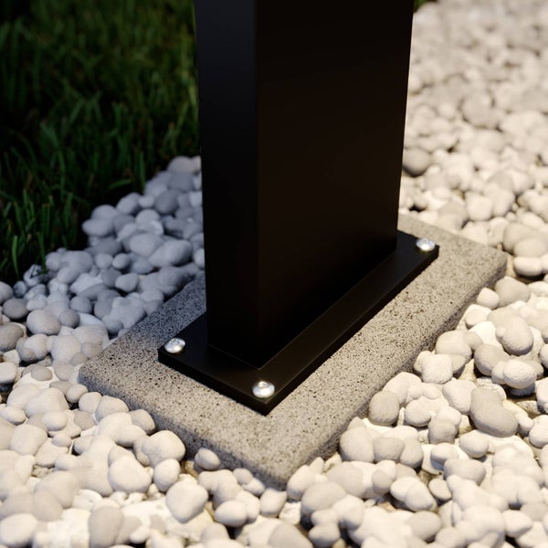 Schwarze Pfostenhalterung aus Metall auf einer Betonplatte, verschraubt und umgeben von weißen Ziersteinen und grünem Gras.