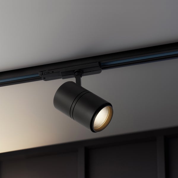 Schwarzer, verstellbarer Schienenstrahler an einer Deckenschiene montiert, strahlt warmweißes Licht aus.