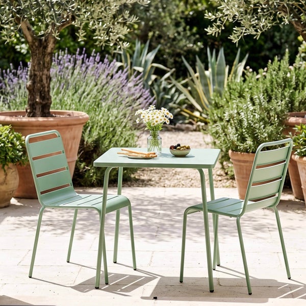 Modernes Gartenset aus Metall mit quadratischem Tisch und zwei Lattenstühlen auf einer hellen Terrasse inmitten mediterraner Bepflanzung.