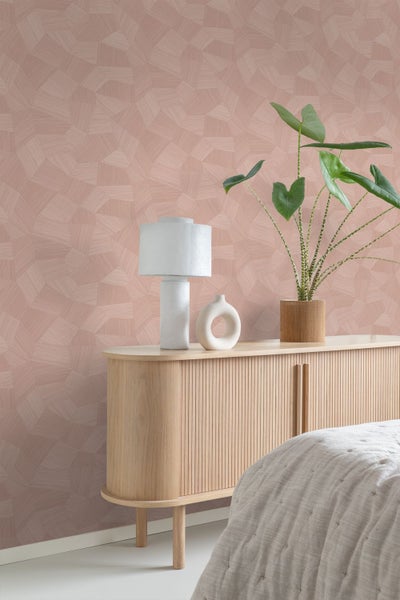 Vliestapete mit geometrischem Mosaikmuster in einem hellen Schlafzimmer hinter einem Sideboard aus Holz mit Lampe und Pflanze.