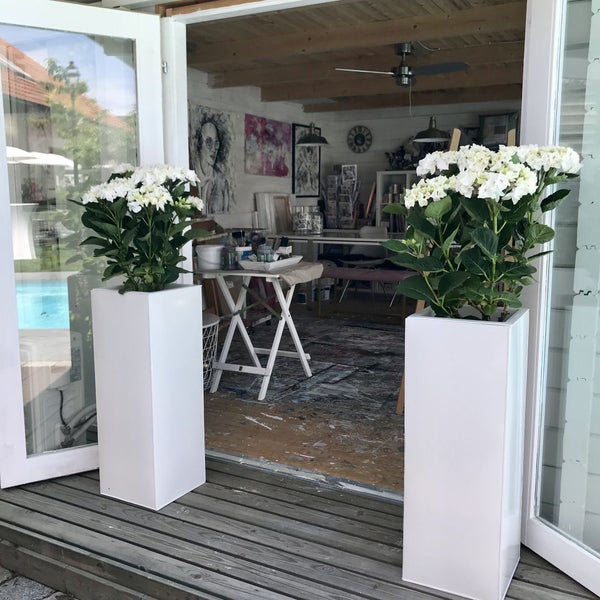 Zwei hohe weiße quadratische Pflanzkübel mit weißen Hortensien auf einer Holzterrasse vor einem hellen Kunstatelier mit Deckenventilator.