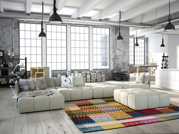 Großes, offenes Wohnzimmer im Loftstil mit hohen Fenstern, Betonwänden und einem modernen beigen Ecksofa auf einem gemusterten bunten Teppich.