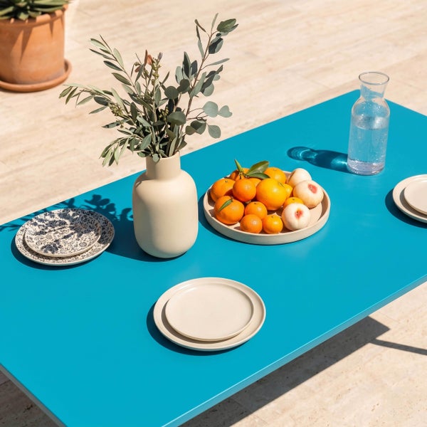 Moderner türkisfarbener Gartentisch gedeckt mit Keramikvase, Obstteller, gemusterten Tellern und Wasserkaraffe auf einer sonnigen Terrasse.