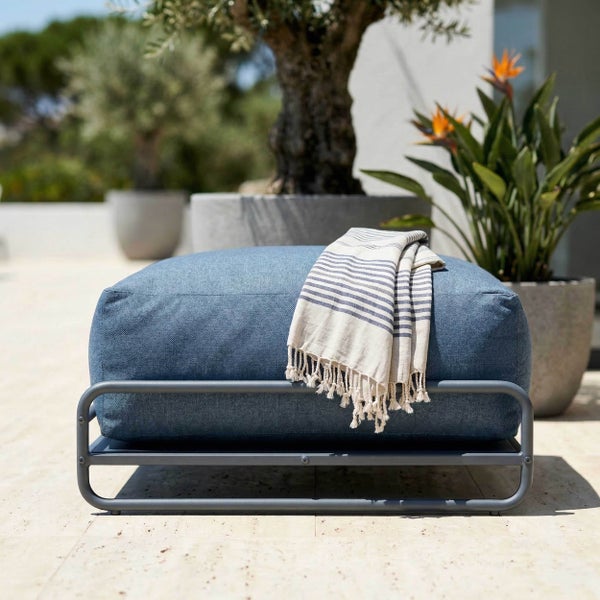 Blauer Outdoor-Hocker auf grauem Metallgestell mit gestreiftem Handtuch auf einer hellen Terrasse neben Topfpflanzen.
