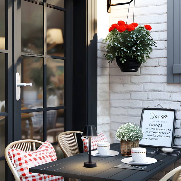 Moderne LED-Tischleuchte auf dunklem Tisch mit Stühlen, karierten Kissen, Kaffeetassen und Blumenampel an einer weißen Ziegelwand.