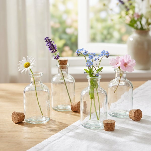 Vier kleine, transparente Glasflaschen mit Korken, dekoriert mit einzelnen Blumen auf einem Holztisch vor einem Fenster.