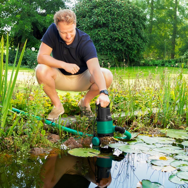 Ein Mann setzt eine grün-schwarze Tauchpumpe in einen hellen Gartenteich mit Seerosen ein. Ein grüner Schlauch ist an der Pumpe befestigt.