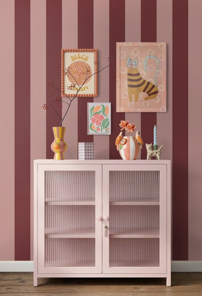Rosa Sideboard mit Glastüren aus Riffelglas vor einer gestreiften Wand, dekoriert mit farbenfrohen Vasen und gerahmter Wandkunst.