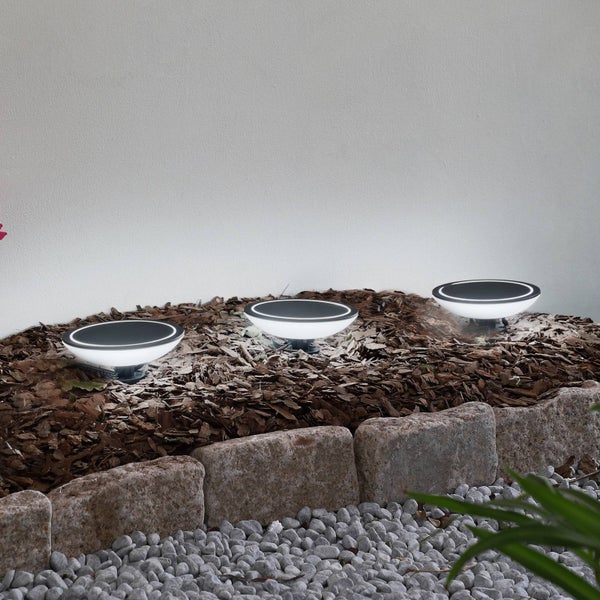 Drei runde LED-Solarleuchten in einem Gartenbeet mit Rindenmulch, eingefasst von Natursteinen und Kieselsteinen.