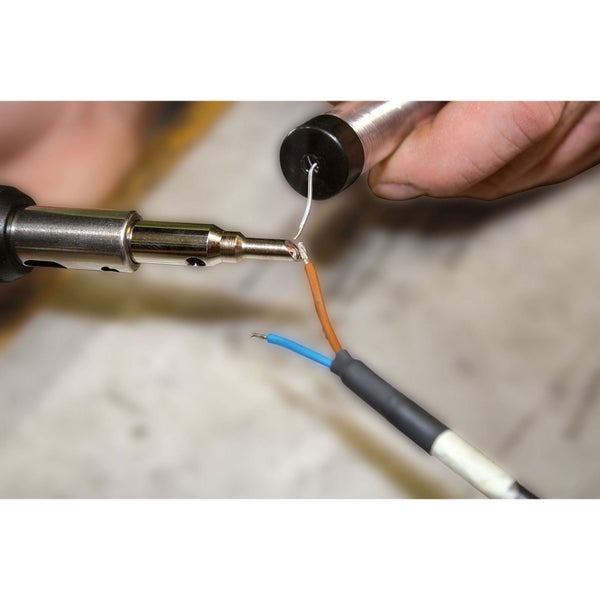 Nahaufnahme einer Hand beim Löten eines elektrischen Kabels mit einem Lötkolben und Lötdraht auf einer Werkbank.