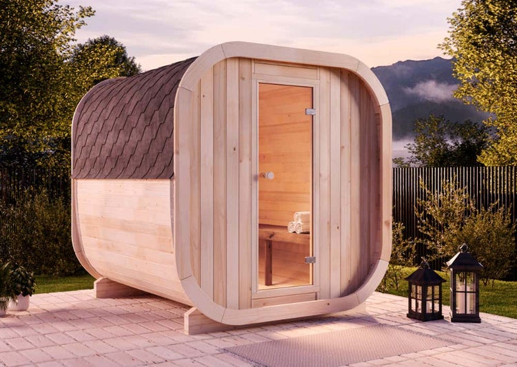 Moderne Fasssauna aus hellem Holz mit Glastür und dunklem Schindeldach, auf einer Terrasse im Garten platziert.