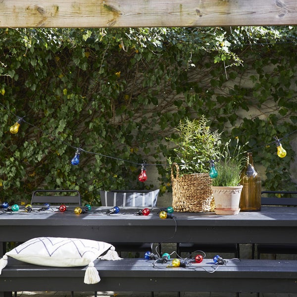 Szene einer Gartenterrasse mit Tisch, Bank, Kissen, Pflanzen und bunter Lichterkette.