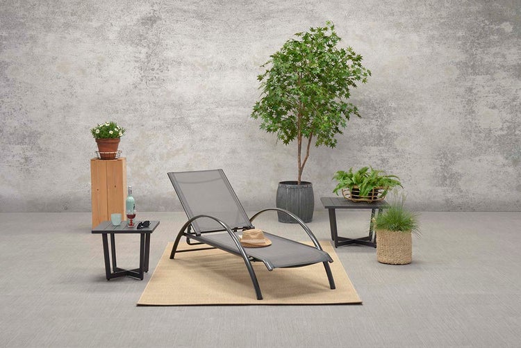 Graue Sonnenliege mit geschwungenen Armlehnen auf einem Teppich, umgeben von Pflanzen und Beistelltischen vor einer strukturierten grauen Wand.
