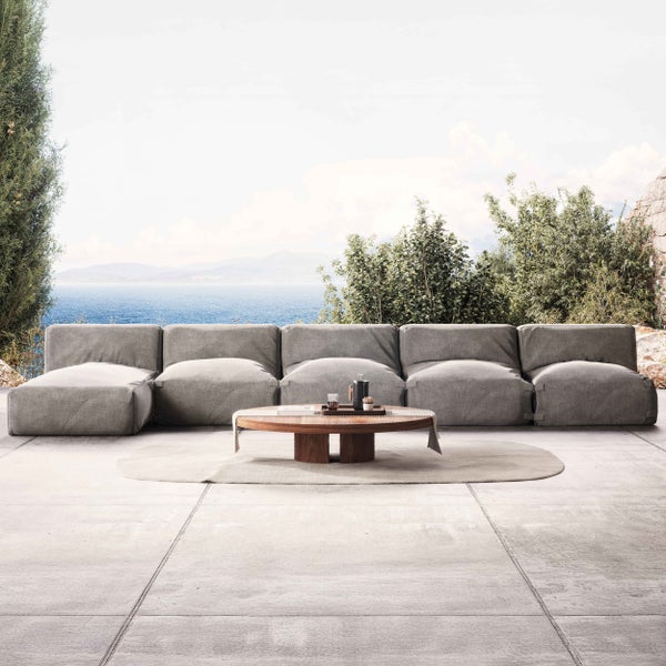 Modulares Gartensofa in Grau auf einer hellen Terrasse mit rundem Holztisch und Meerblick im Hintergrund.