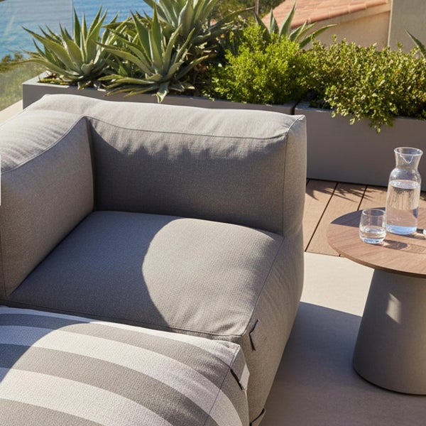 Moderner Outdoor-Loungebereich mit grauem Polstereckmodul, gestreiftem Hocker und kleinem Holzbeistelltisch auf einer Terrasse mit Pflanzen.