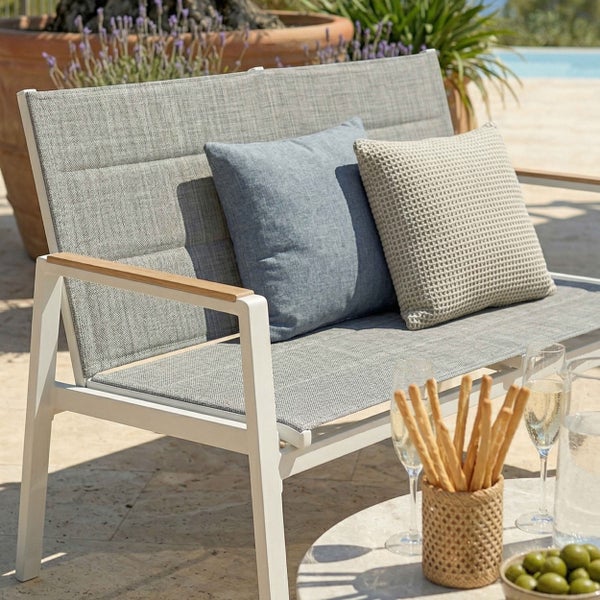 Moderne Gartenbank mit weißem Aluminiumgestell, Holzarmlehnen und grauer Textilbespannung, dekoriert mit Kissen auf einer hellen Steinterrasse.