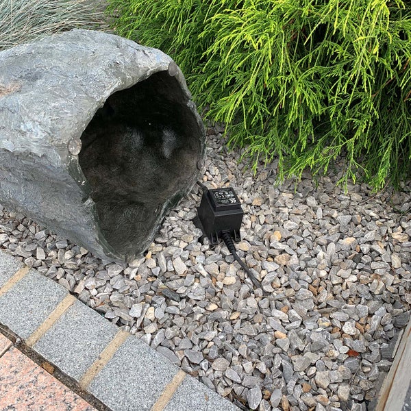 Künstlicher Stein in Steinoptik auf einem Kiesbett neben einem schwarzen Transformator mit Kabel vor einer grünen Gartenpflanze.