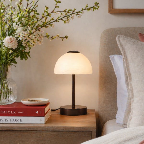 Kleine Tischlampe mit halbkugelförmigem Glasschirm auf einem Nachttisch neben Blumen und Büchern in einem Schlafzimmer.