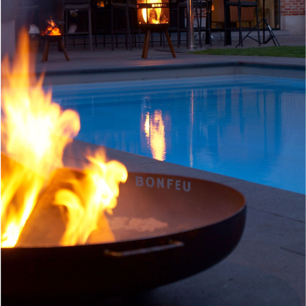 Moderne schwarze BonFeu Feuerschale aus Metall mit Flammen auf einer Steinterrasse am Swimmingpool im Garten.