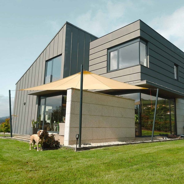 Modernes Haus mit anthrazitfarbener Fassade und beigem dreieckigem Sonnensegel, das an schwarzen Pfosten im Garten befestigt ist.