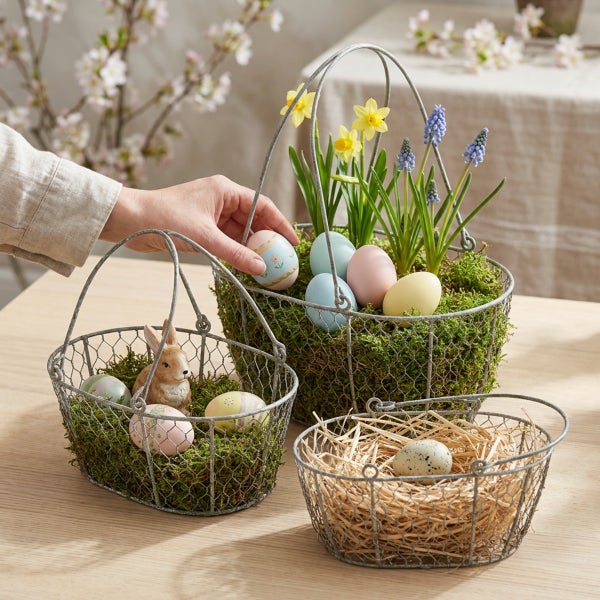 Drei rustikale Drahtkörbe zur Osterdekoration, gefüllt mit Moos, Ostereiern, einer Osterhasenfigur und Frühlingsblumen auf einem Holztisch.