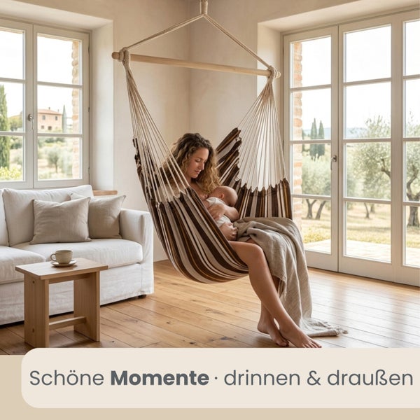 Eine Frau mit Baby sitzt in einem gestreiften Hängesessel in einem hellen Wohnzimmer mit Holzfußboden und großen Fenstern.