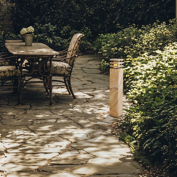 Beleuchteter Sandstein Wegeleuchte im Garten neben einem rustikalen Holztisch und Blumenbeeten auf einer Natursteinpflaster Terrasse