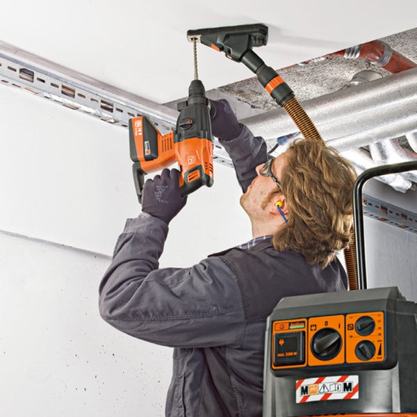 Handwerker bohrt mit einem Akku-Bohrhammer in eine Decke, während ein Staubsauger den Bohrstaub über einen Adapter direkt absaugt.