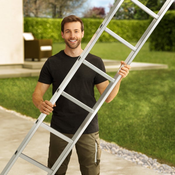 Lächelnder Handwerker hält eine Aluminium Anlegeleiter im Garten vor einem Haus.
