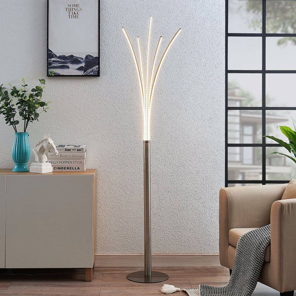 Moderne LED-Stehleuchte aus Metall mit fünf gebogenen Lichtstäben in einem hellen Wohnzimmer neben einem Sessel und Sideboard.