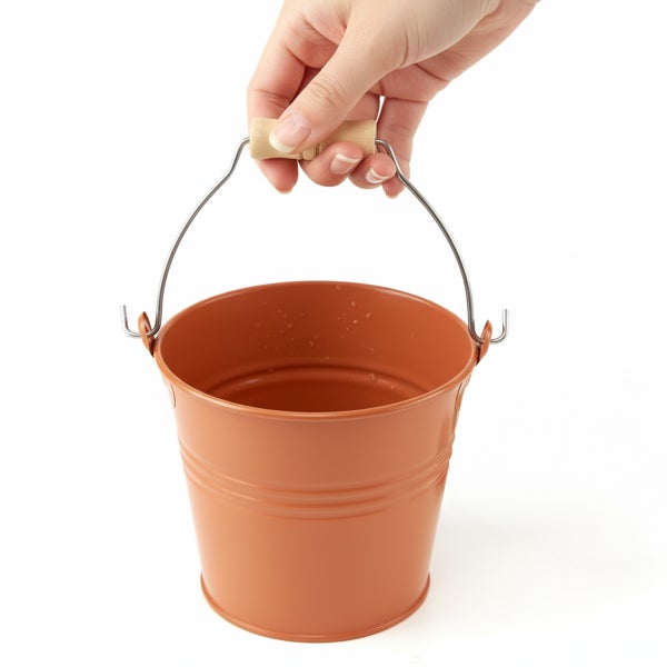 Kleiner terrakottafarbener Metalleimer mit Holzgriff am Henkel, von einer Hand gehalten.