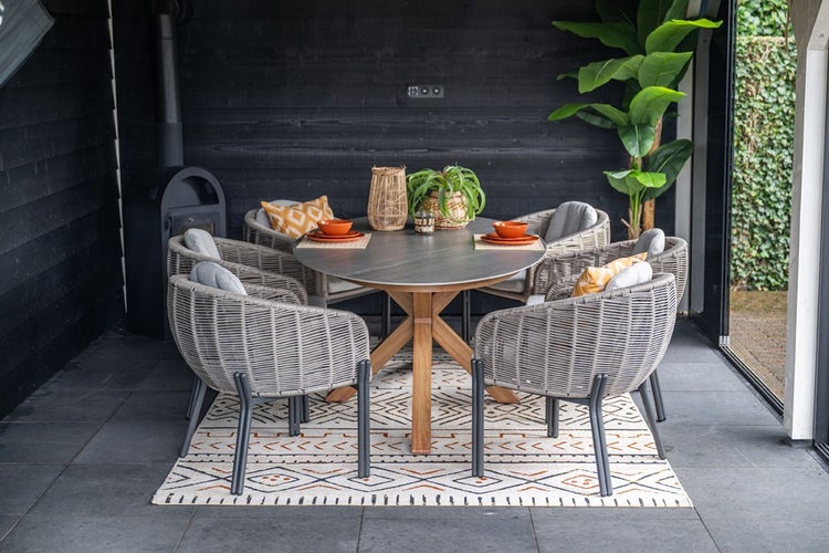 Moderne Garten-Essgruppe mit rundem Holztisch und grauer Platte sowie grauen Sesseln in Korboptik mit Polstern auf einem Teppich.