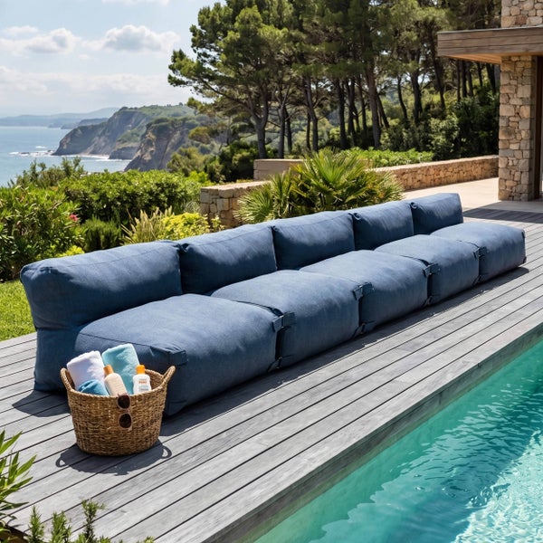Modulares Outdoor-Lounge-Sofa auf einer Holzterrasse am Pool mit Meerblick, große Stoffkissen und ein geflochtener Korb.