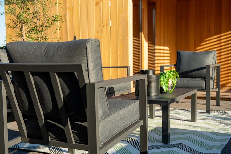 Lounge-Set mit zwei Metall-Sesseln, grauen Polstern und Couchtisch auf einem gemusterten Outdoor-Teppich vor einer hellen Holzwand.