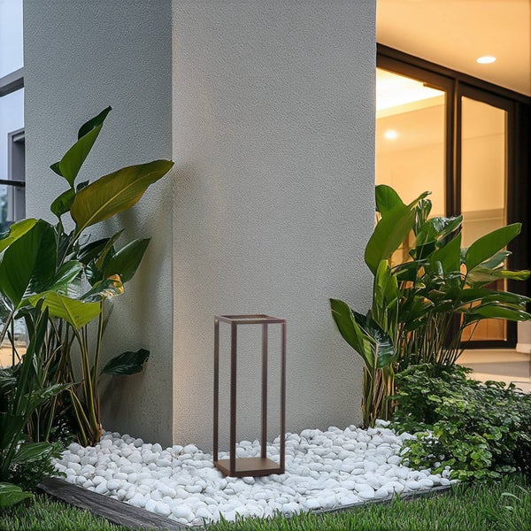 Moderne rechteckige Außenleuchte auf weißen Kieseln in einem Garten neben großen grünen Pflanzen an einer strukturierten grauen Wand.