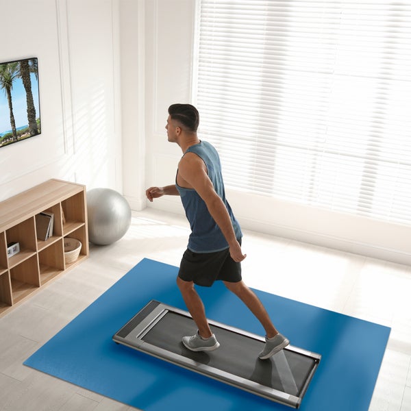 Mann beim Training auf einem flachen Laufband in einem hellen Raum. Das Fitnessgerät steht auf einer Bodenschutzmatte.