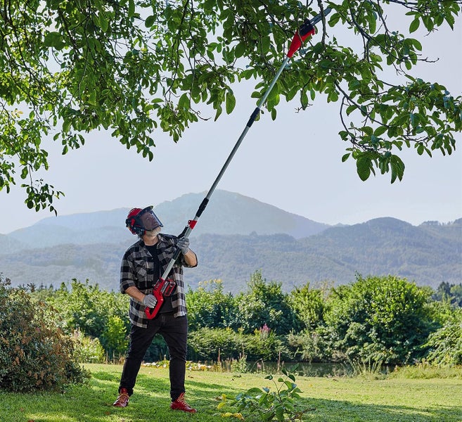 Person nutzt einen Teleskop-Hochentaster zur Baumpflege in einem Garten vor einer Bergkulisse.