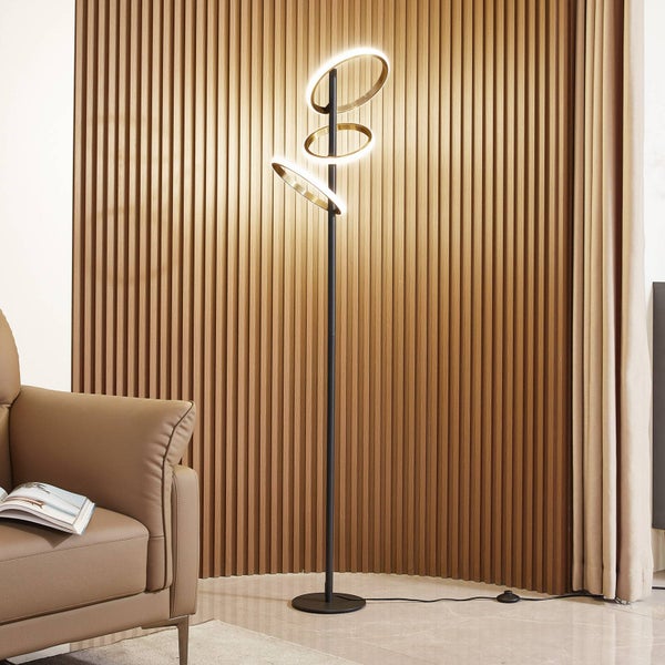 Moderne LED-Stehleuchte mit drei ringförmigen Elementen, schwarzes Gestell, vor einer Wand mit Holzpaneelen neben einem Ledersessel.